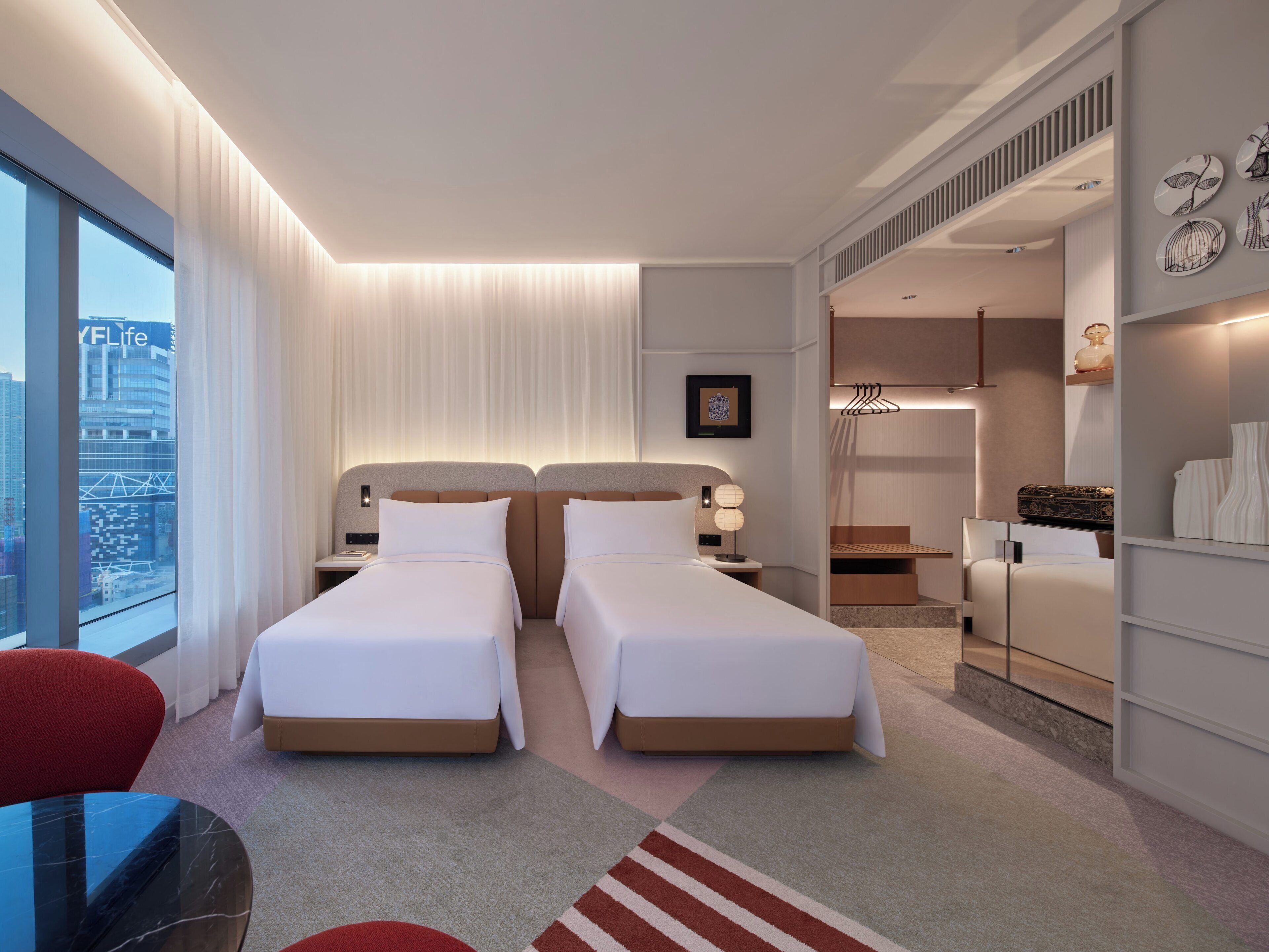 A modern hotel room with two beds, a sitting area with a table and chairs, a view of the city, and a walk-in closet.