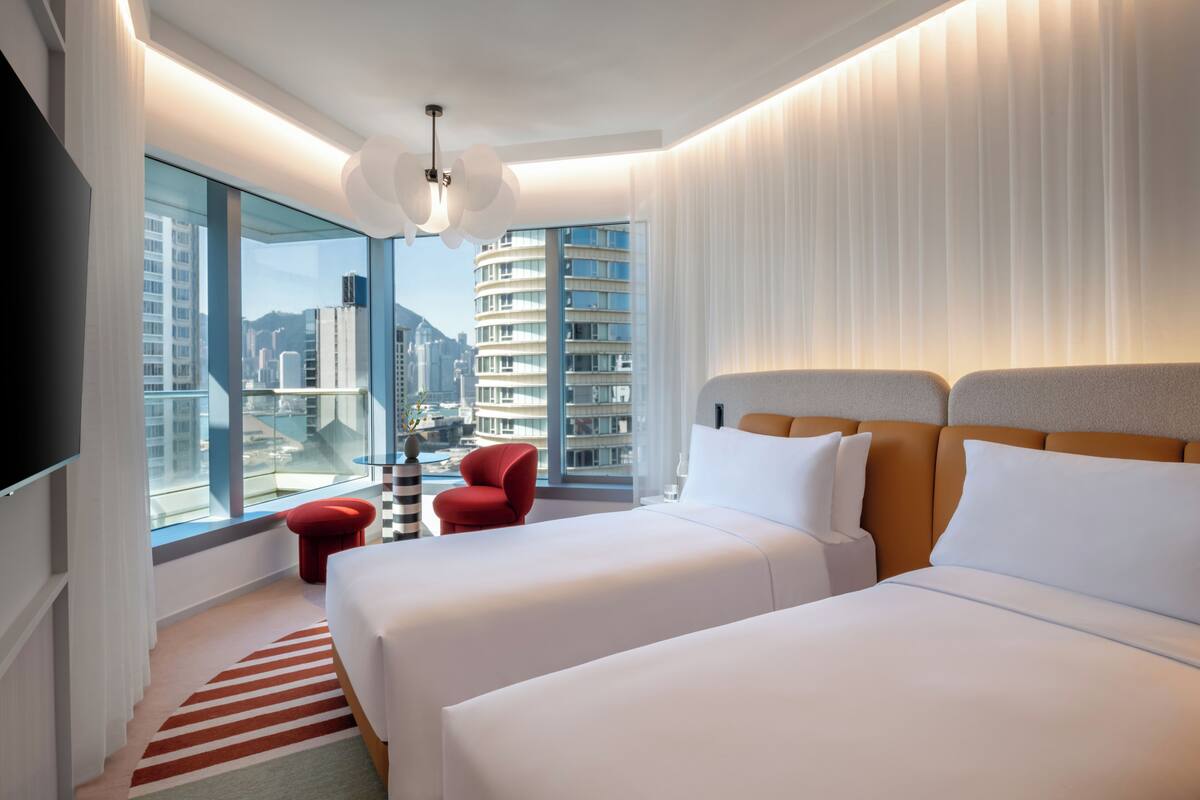 A modern hotel room with two beds, a red chair, a large window with a city view, and a striped rug.