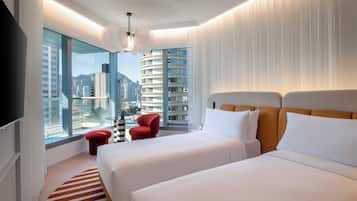 Quarto de hotel moderno com duas camas, uma cadeira vermelha, uma janela grande com vista para a cidade e um tapete listrado.