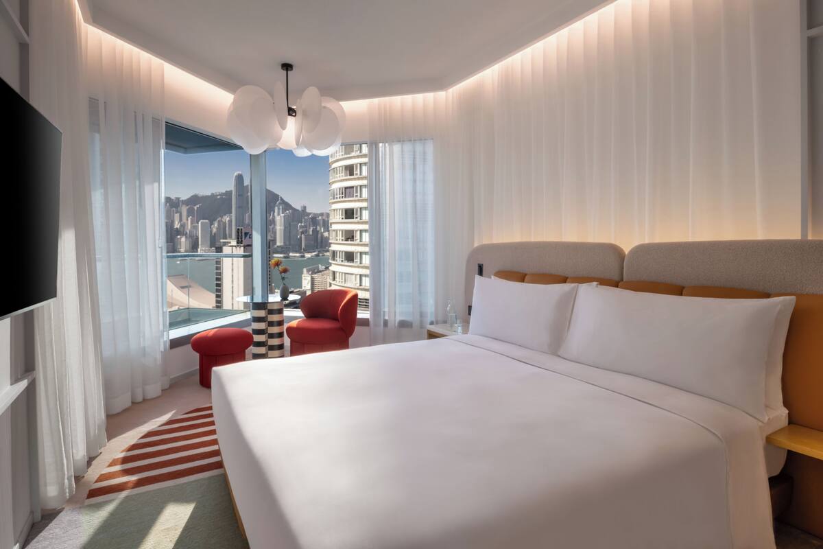 A hotel room with a large bed, a red chair, a TV, and a view of a cityscape.