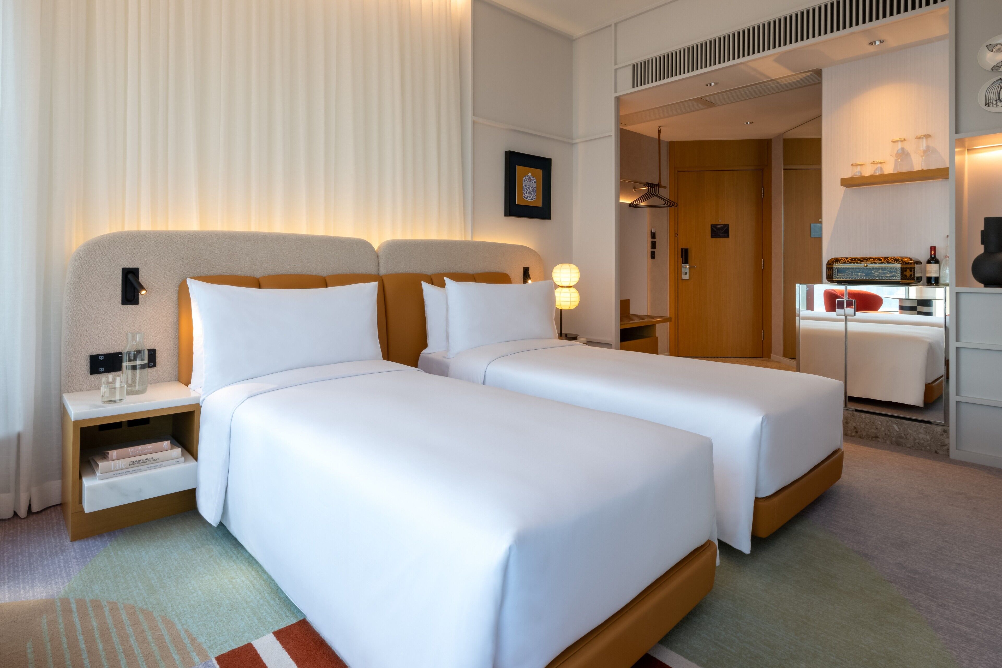 A hotel room with two beds, a bedside table with a lamp, a wardrobe, and a small seating area with a table and chairs.