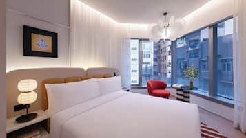 A modern hotel room with a large bed, a red chair, a side table with a vase, and a view of the city through large windows.
