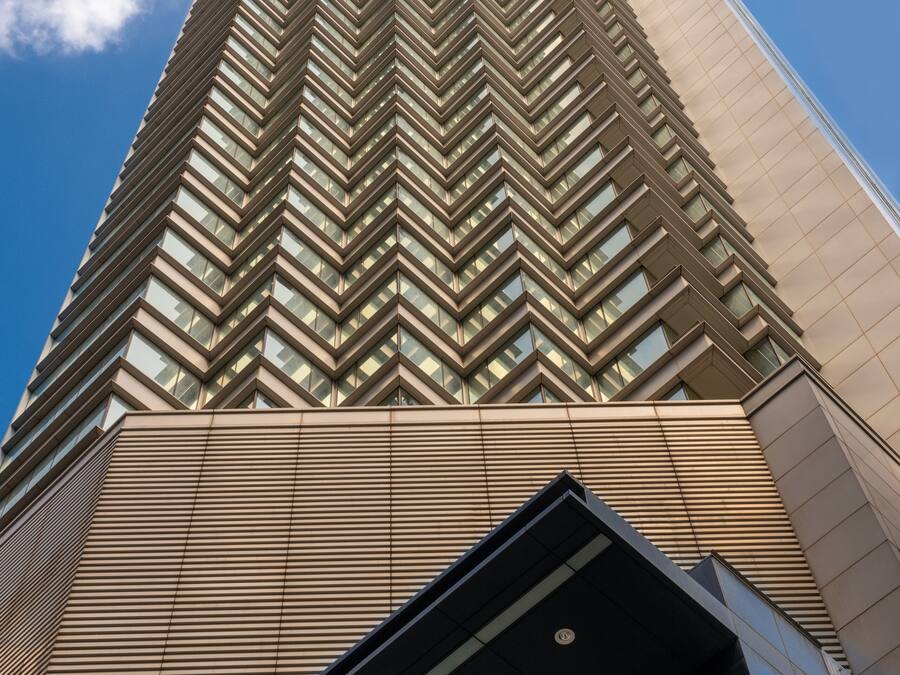 A tall, modern building with a zigzag pattern of windows and a prominent entrance with glass doors.