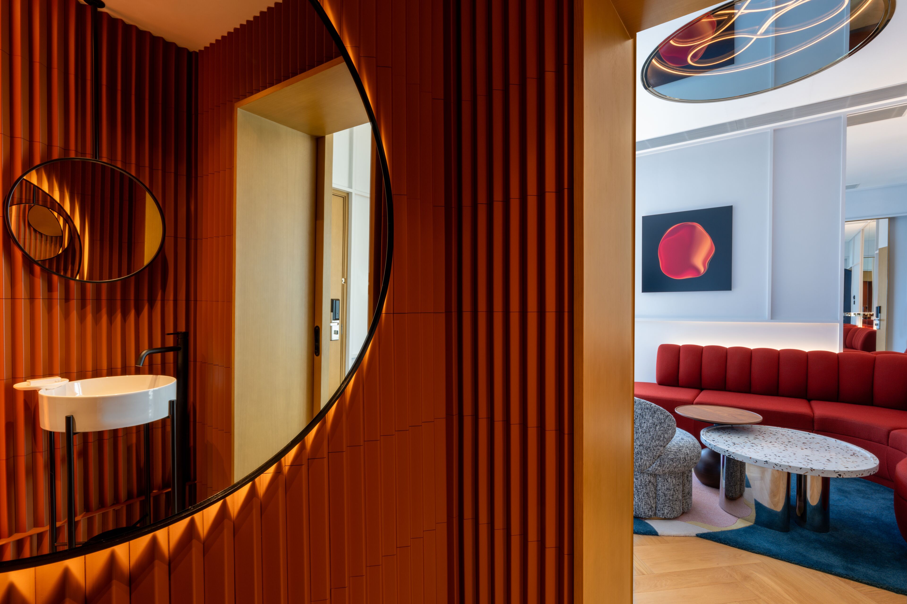 Un interior moderno con una pared de madera, un espejo redondo, un lavamanos blanco, un sofá rojo y una mesa redonda.