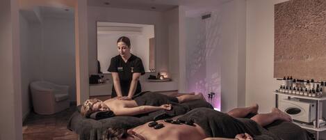 Ein Spa-Raum mit zwei Klienten, die eine Massage erhalten, einem Massagetisch, einer Massagebehandlerin und einer Ablage mit Massageölen.