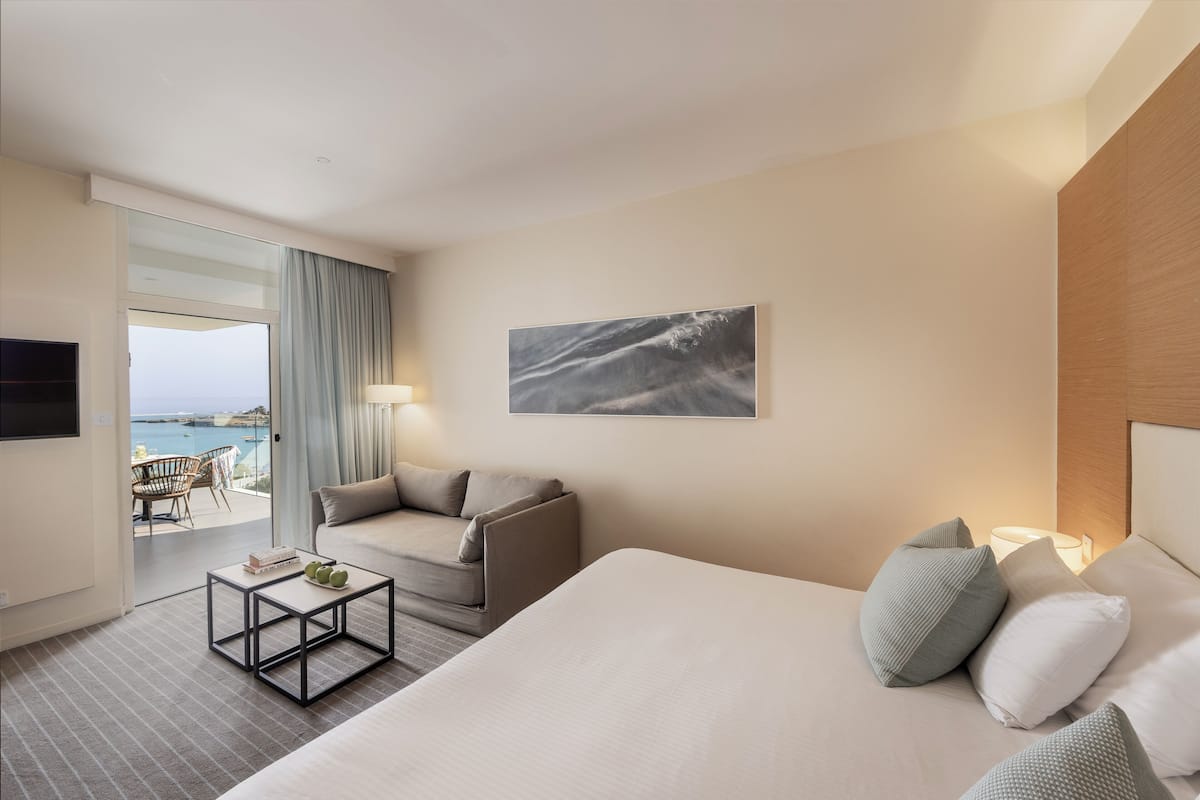 A hotel room with a bed, a sofa, a small table, and a view of the sea through a glass door.