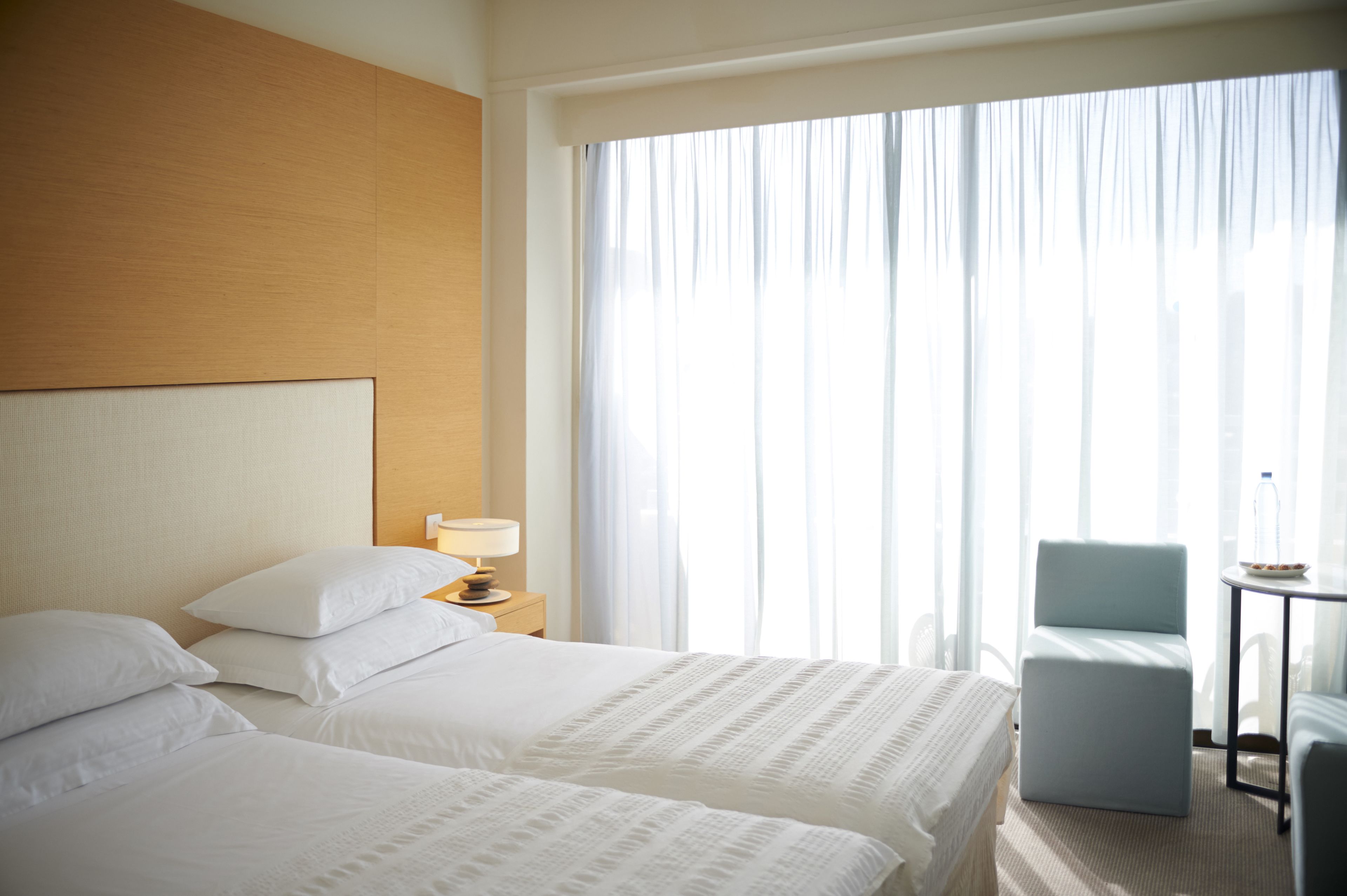 Een hotelkamer met twee bedden, een nachtkastje met een lamp, een stoel en een kleine tafel met een fles en een schaal.
