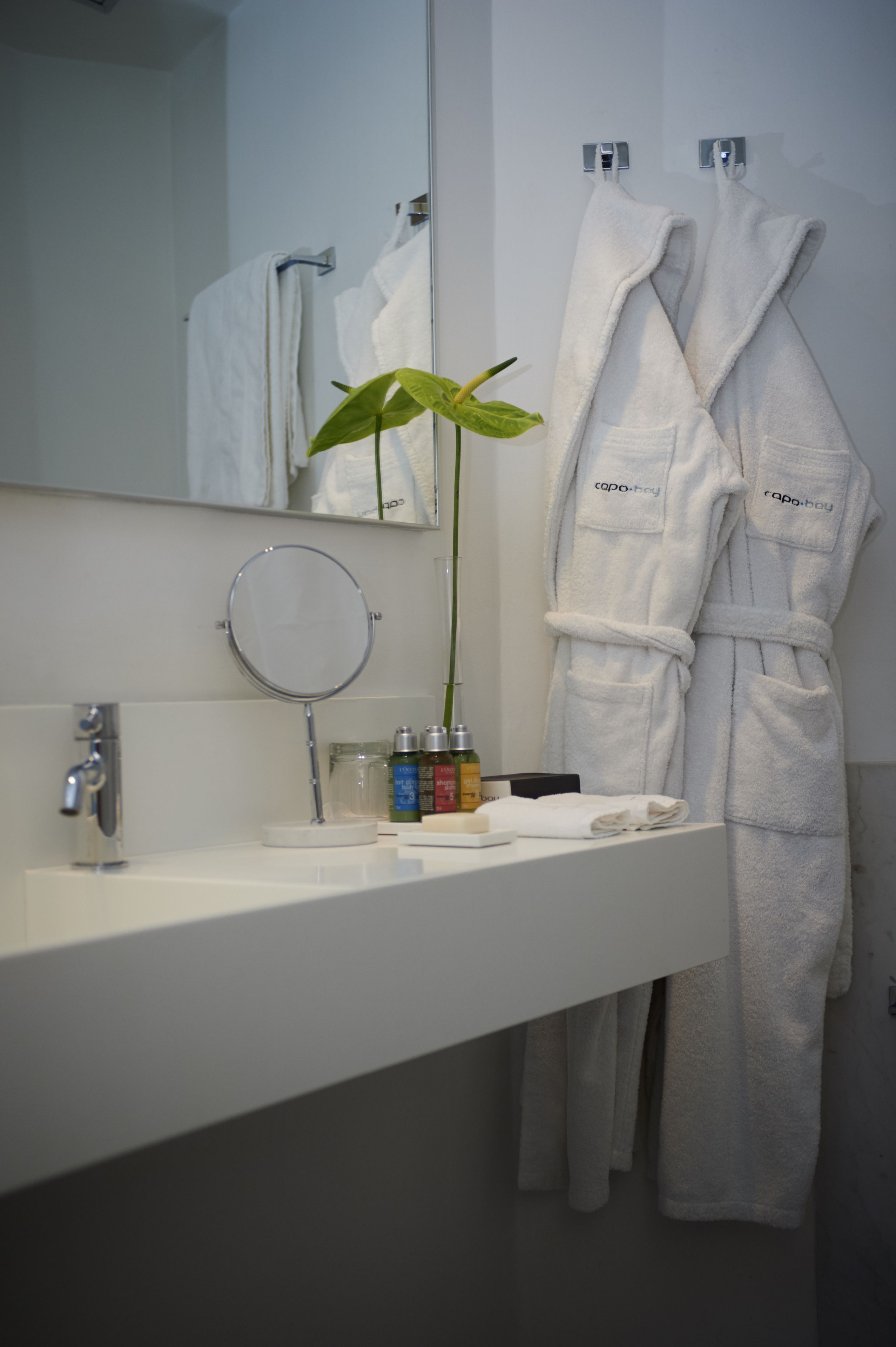 Un baño con un lavabo blanco, un espejo, una planta y dos batas blancas colgadas de ganchos.