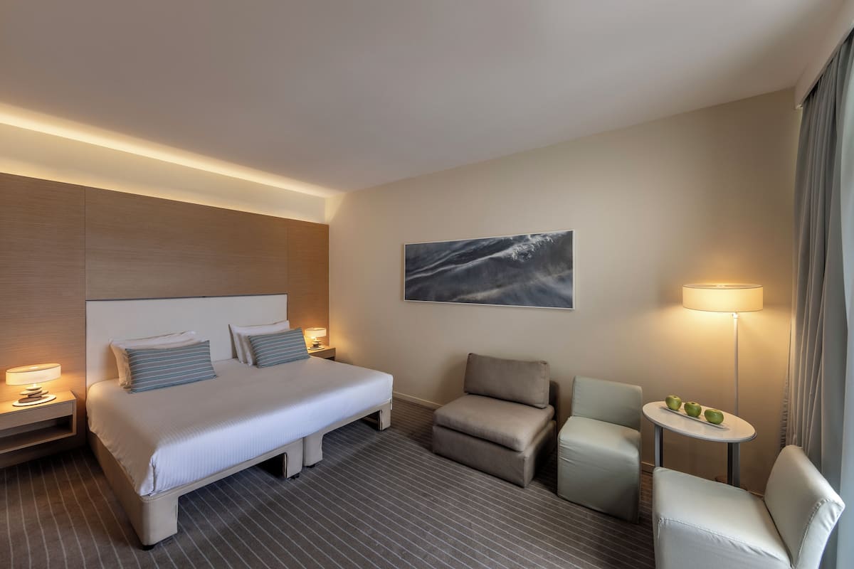 A hotel room with a large bed, two bedside tables with lamps, a small round table with apples, two armchairs, and a painting on the wall.