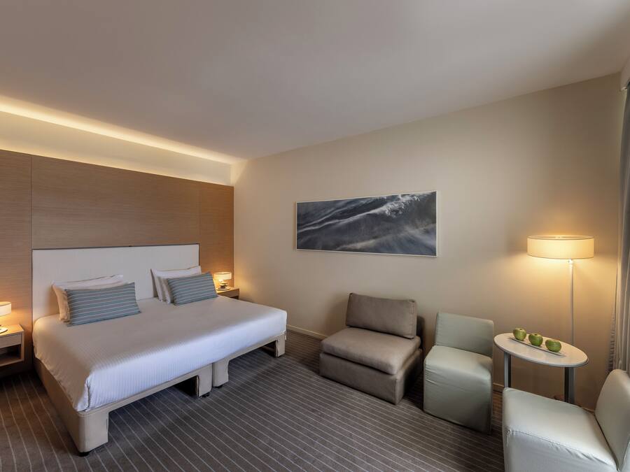 Habitación de hotel con una cama grande, dos mesitas de noche con lámparas, una pequeña mesa redonda con manzanas, dos sillones y un cuadro en la pared.