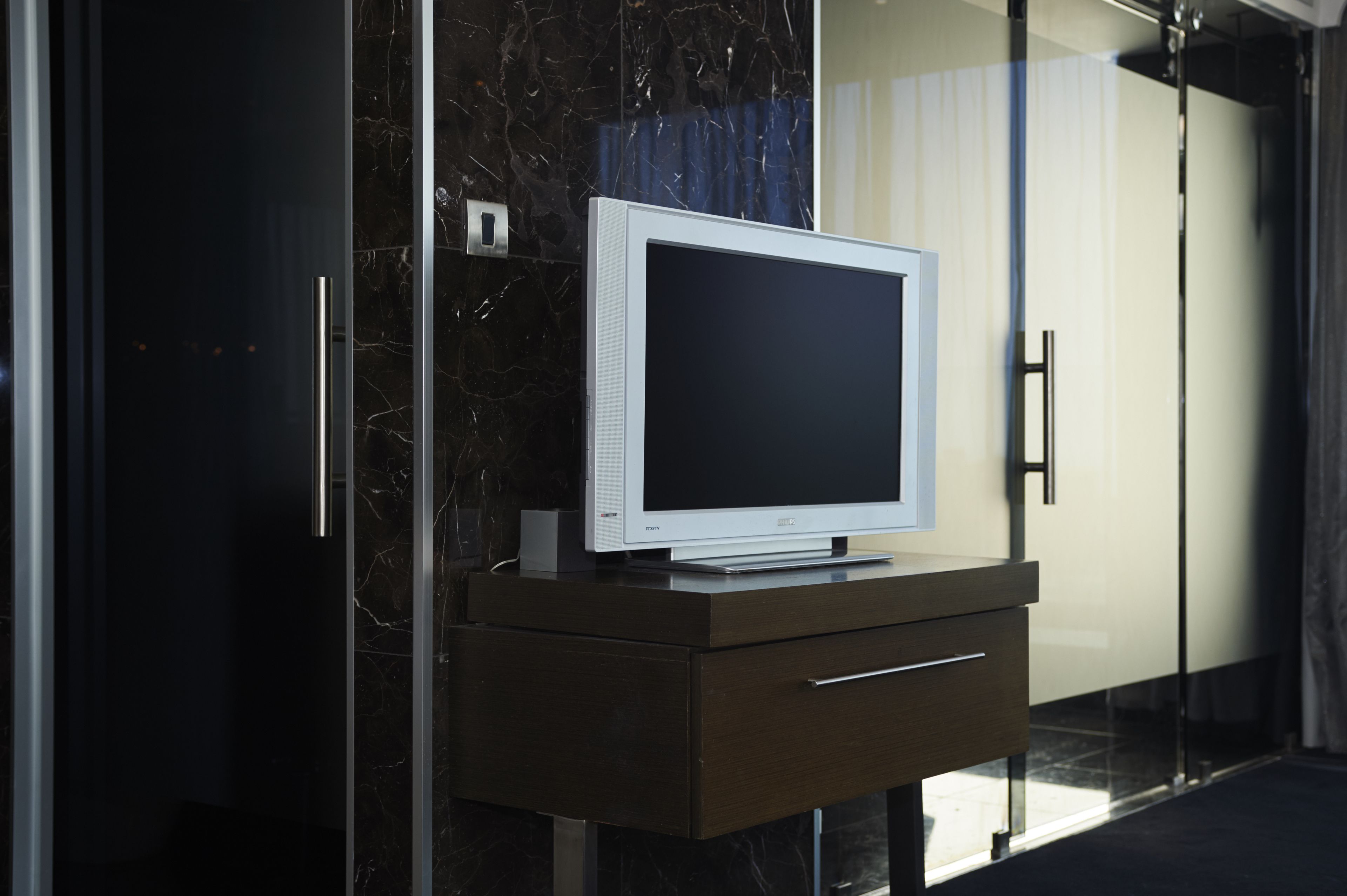Un baño moderno con cabina de ducha de vidrio, pared con azulejos de mármol y tocador blanco con televisor de pantalla plana.