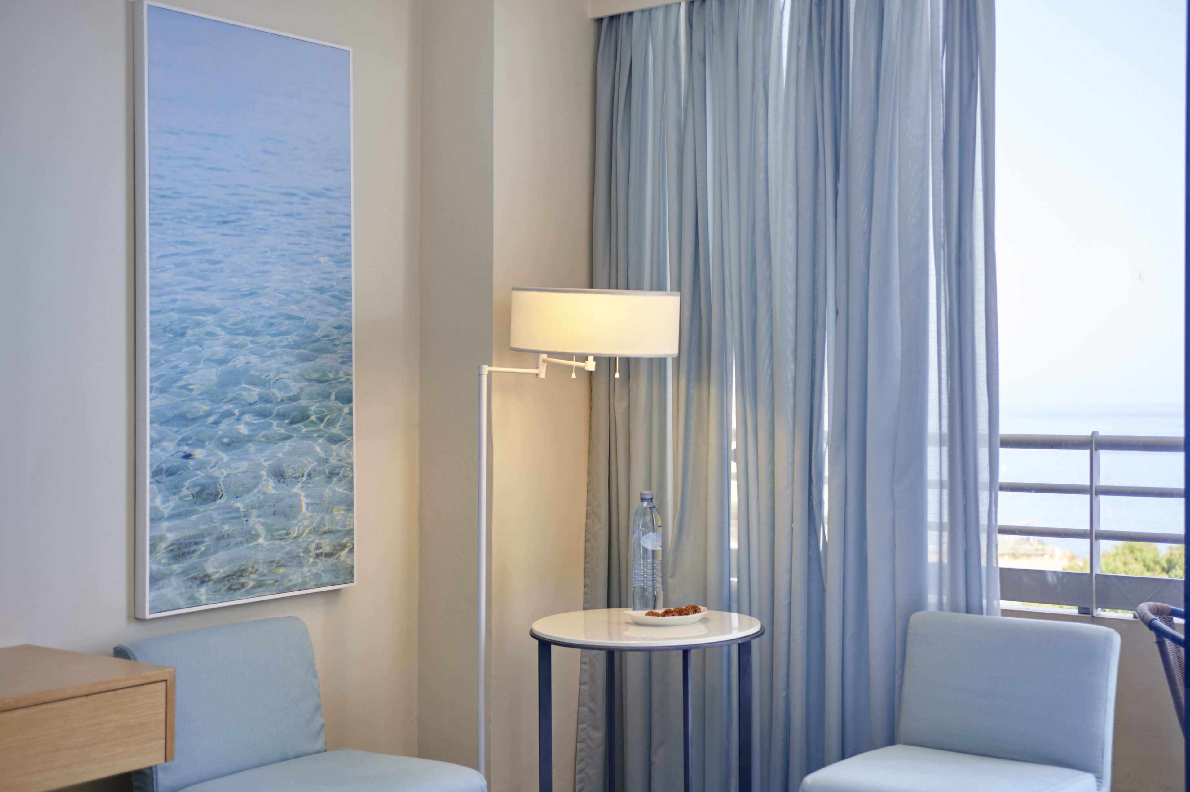Una camera d'albergo con una grande finestra, un tavolo con una lampada, una sedia e un quadro raffigurante una scena marina.