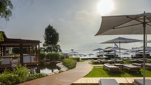 A wooden deck leading to a pool area with lounge chairs and umbrellas, surrounded by greenery and a view of the water. - Capo Bay Hotel (Paralimni)