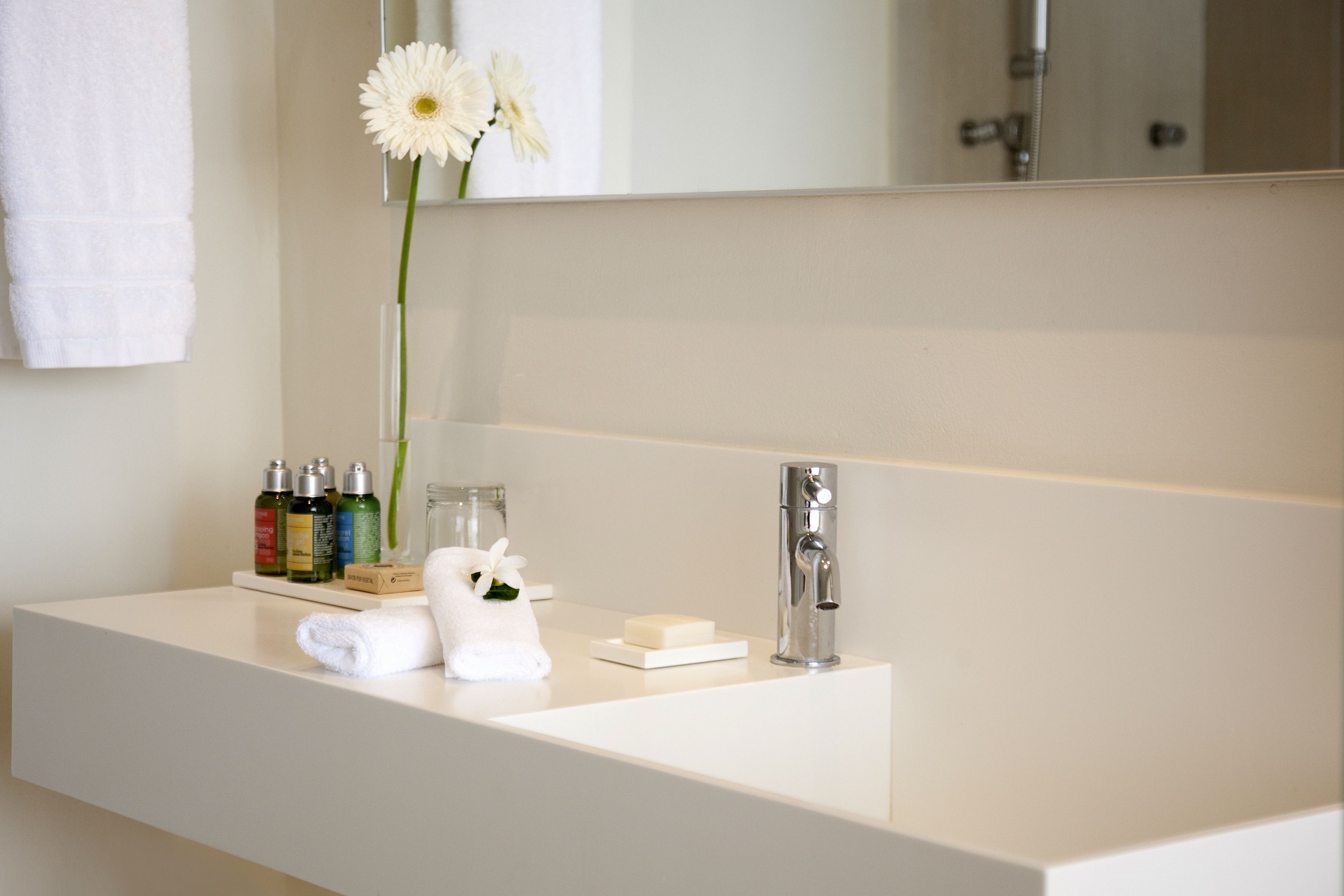 A modern bathroom with a white vanity, a mirror, a towel, a flower, and toiletries.