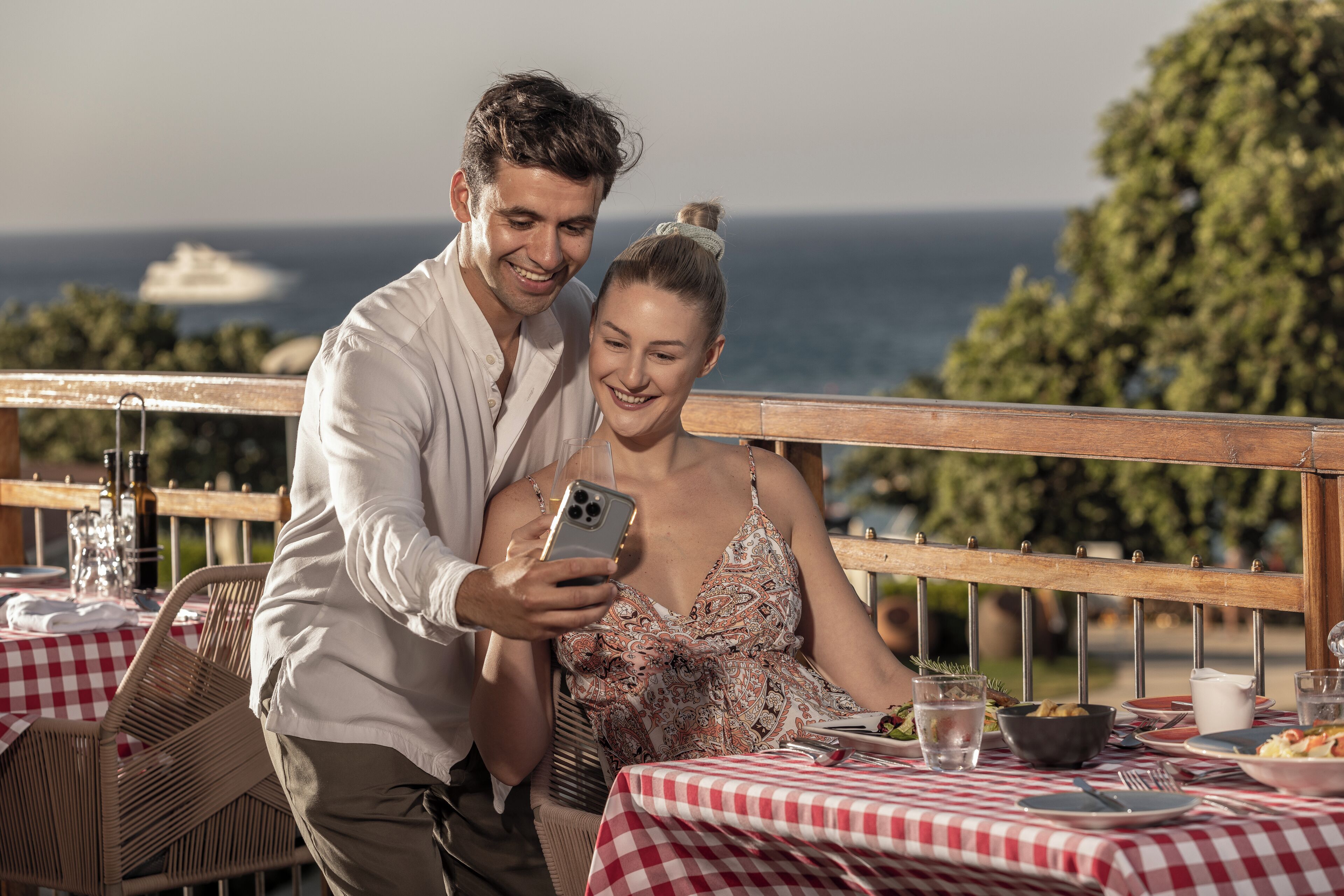 Una pareja cenando en una mesa al aire libre, con vista al mar y un barco en el fondo.
