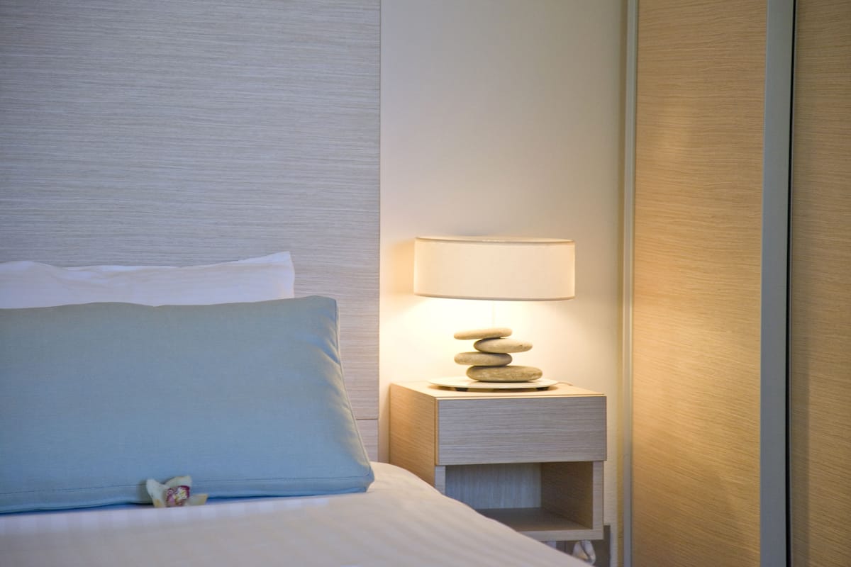 A neatly made bed with a blue pillow and a small decorative item, a bedside table with a lamp and a stack of stones, and a wooden panel wall.