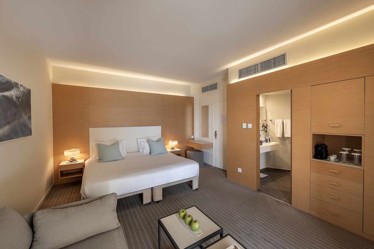 A modern hotel room with a large bed, bedside tables, a sofa, a small table with apples, a mirror, and a wardrobe.