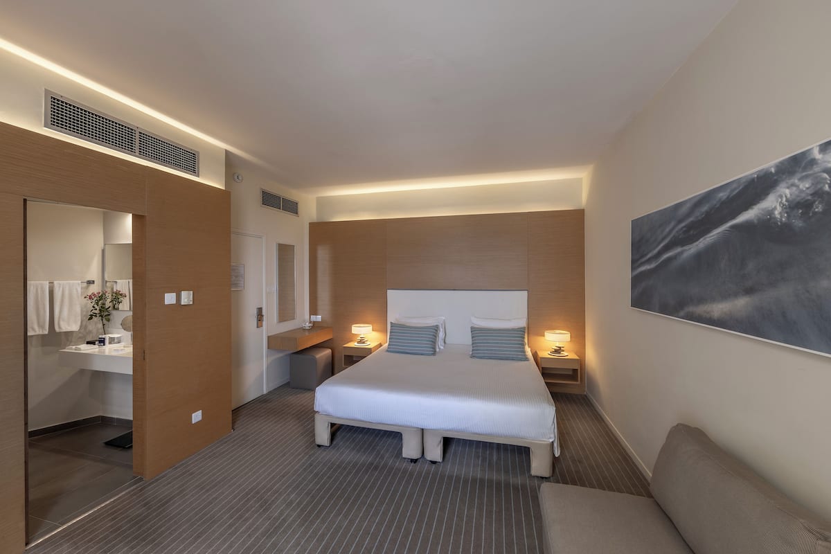 A modern hotel room with a large bed, bedside tables with lamps, a bathroom with a mirror and towel rack, and a painting on the wall.