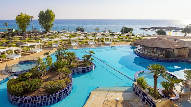 A resort pool area with multiple sections, palm trees, and beach umbrellas. - Capo Bay Hotel (Paralimni)