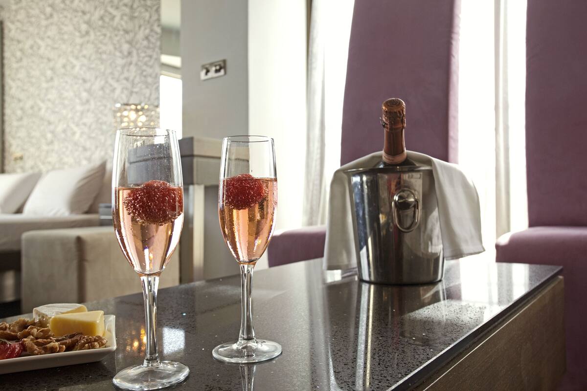 Two champagne flutes with a red garnish on a bar counter, a bottle of champagne, and a plate of cheese and nuts.