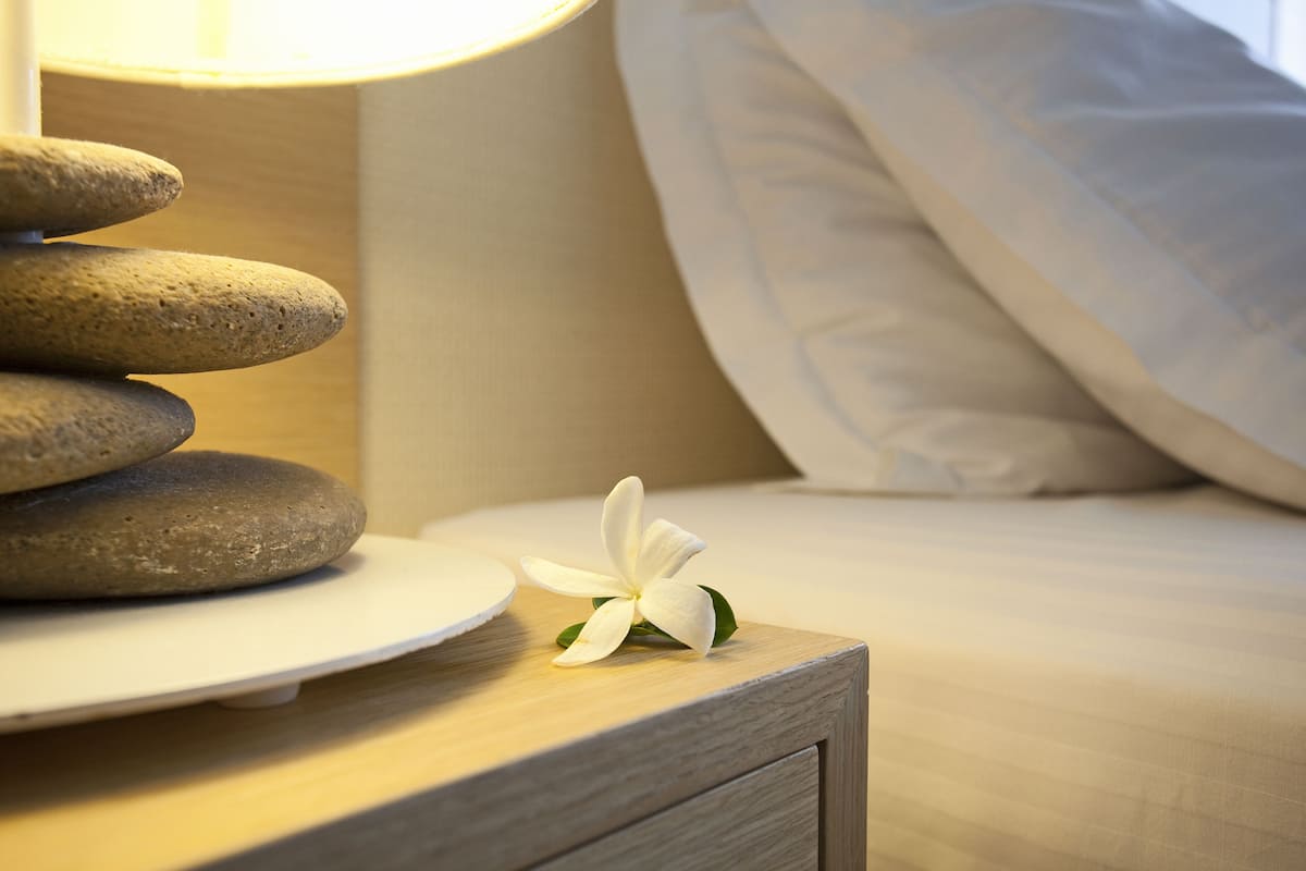 A neatly arranged stack of smooth stones on a wooden nightstand, a white flower, and a bed with white bedding.