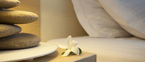 A neatly arranged stack of smooth stones on a wooden nightstand, a white flower, and a bed with white bedding.