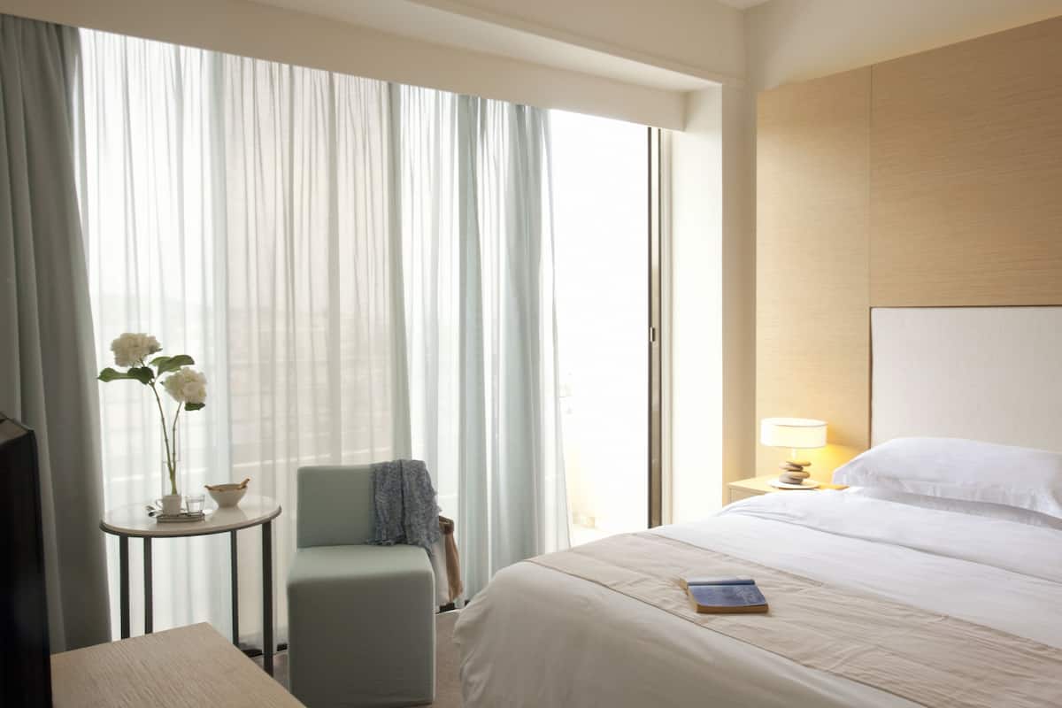 A hotel room with a bed, a chair, a small table with a vase of flowers, and a nightstand with a lamp.
