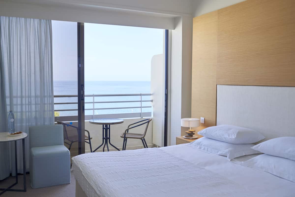 A hotel room with a bed, a small table, two chairs, a nightstand, and a view of the sea.
