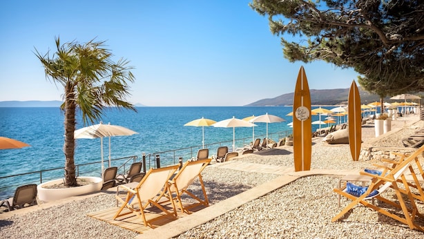 A beachside seating area with lounge chairs and umbrellas, a palm tree, and a surfboard.