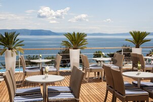 Una terraza con mesas redondas y sillas de mimbre, con vistas a una masa de agua y montañas lejanas.