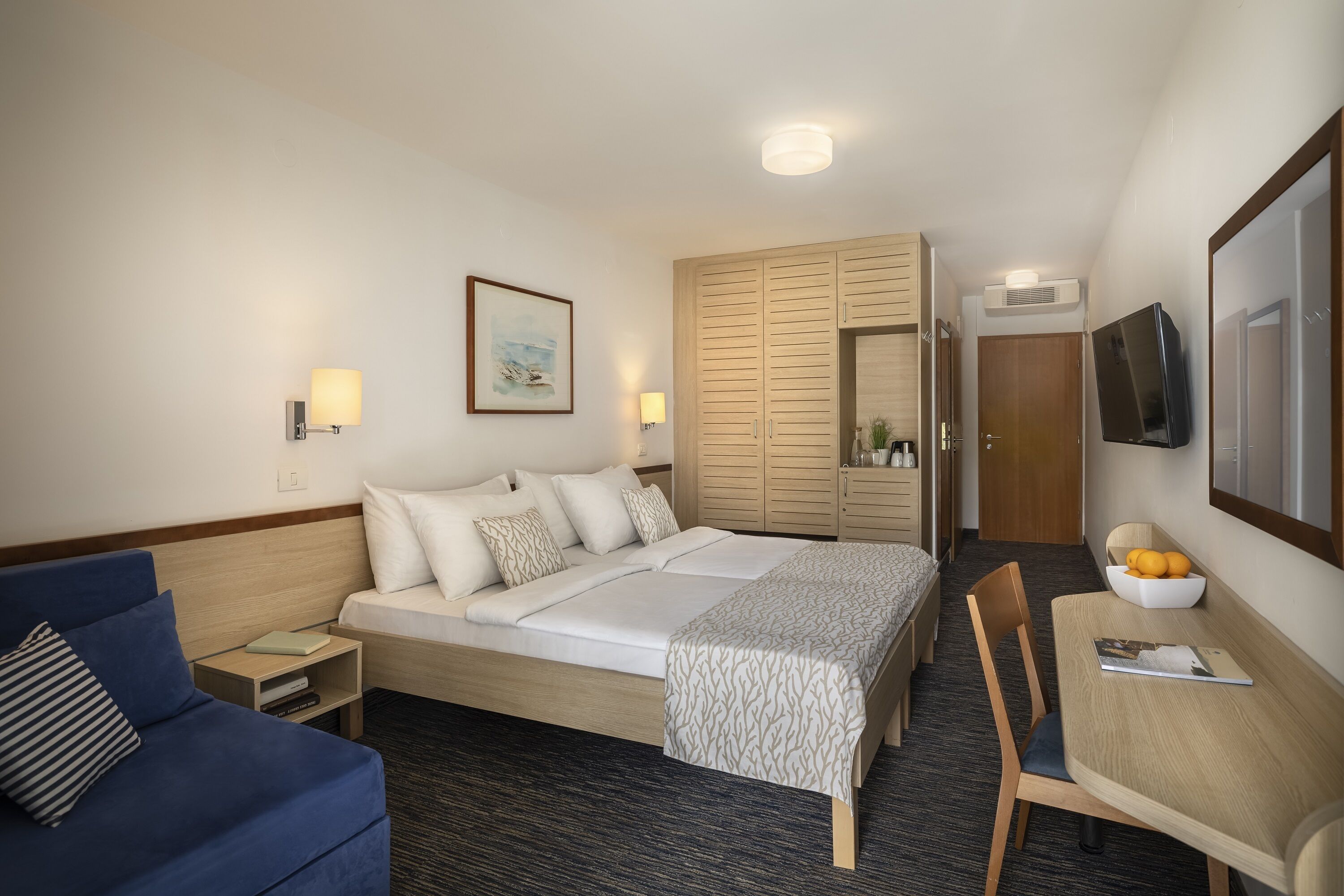 A hotel room with a bed, a blue sofa, a desk with a bowl of fruit, and a TV mounted on the wall.