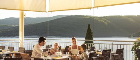 A dining area with tables and chairs, overlooking a body of water and hills.