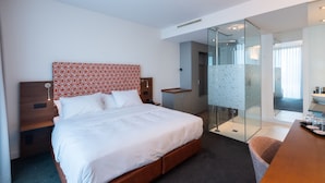 Una habitación de hotel moderna con una cama grande, una mesita de noche, un escritorio y una cabina de ducha de vidrio.