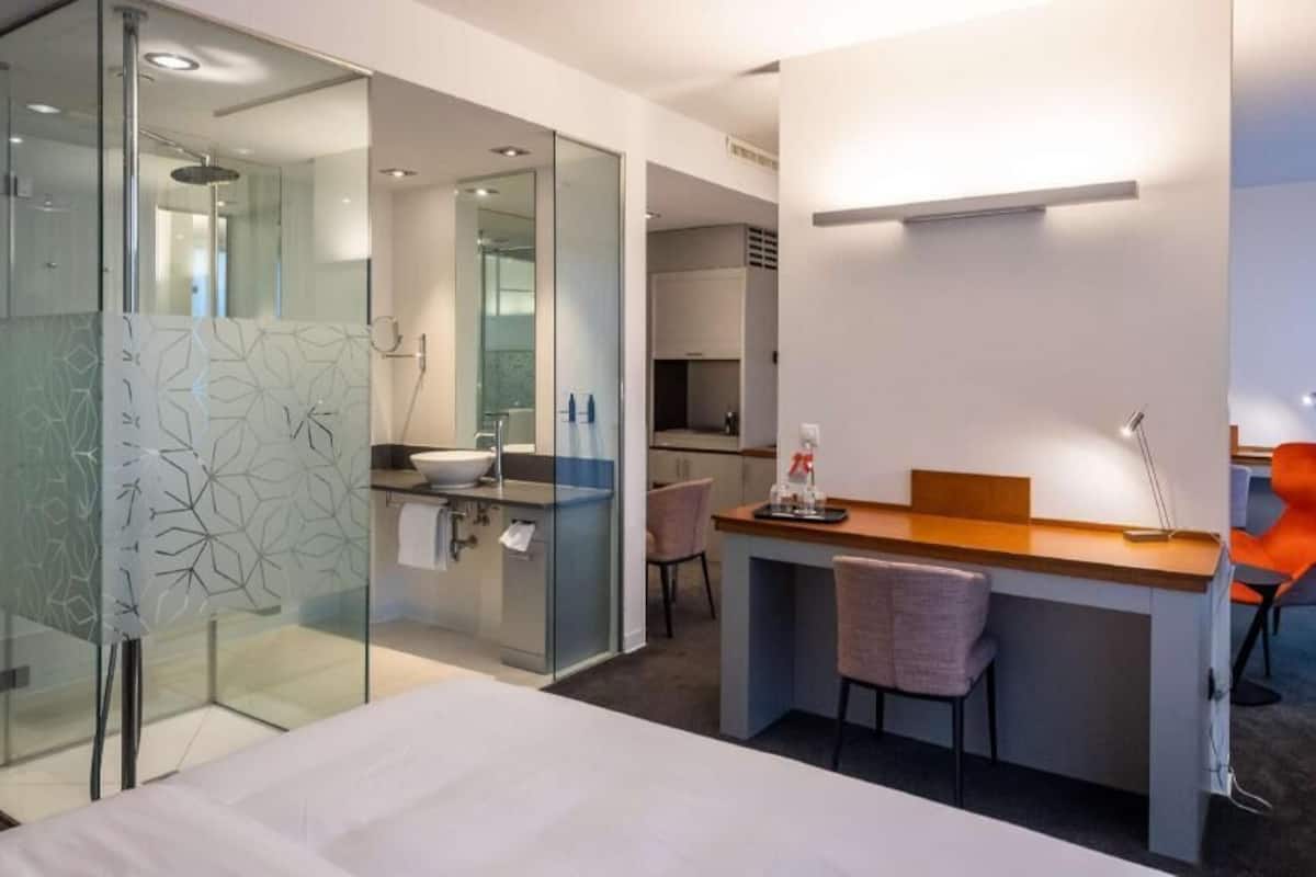 Una habitación de hotel moderna con cabina de ducha de vidrio, un escritorio de madera, dos sillas, una cama y una pequeña kitchenette.