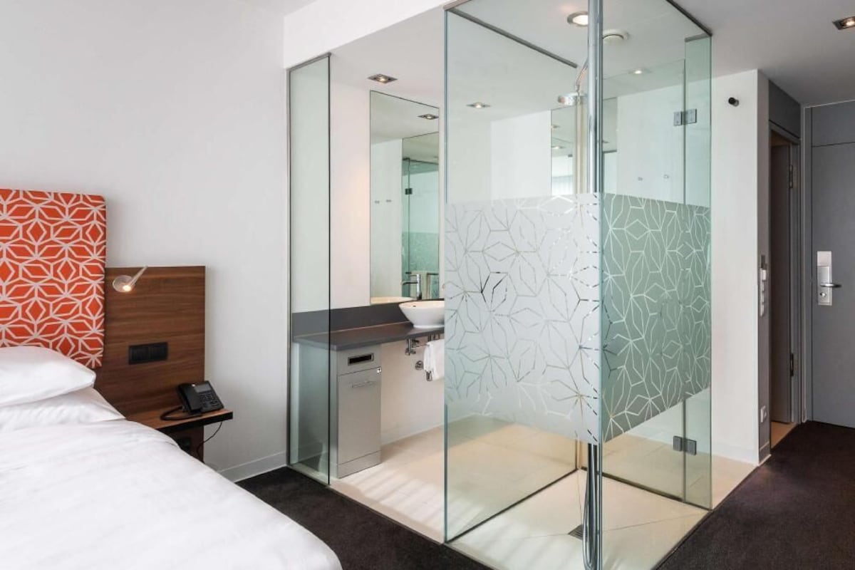 Una habitación de hotel moderna con un separador de vidrio, una cama, una mesita de noche con un teléfono, un baño con un lavamanos y un espejo, y una puerta.