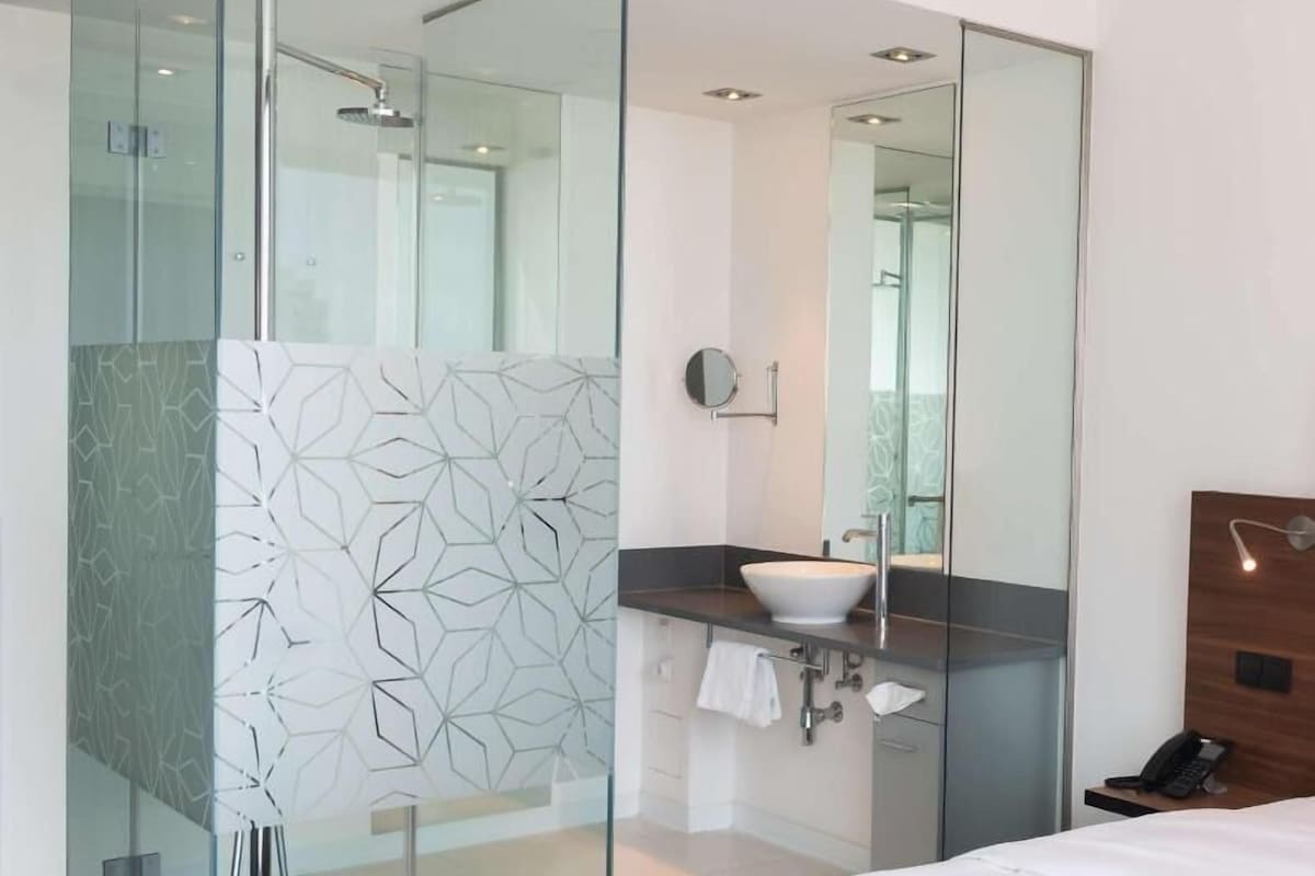 Una habitación de hotel moderna con ducha de vidrio, lavamanos junto a la cama y cabecero de madera.