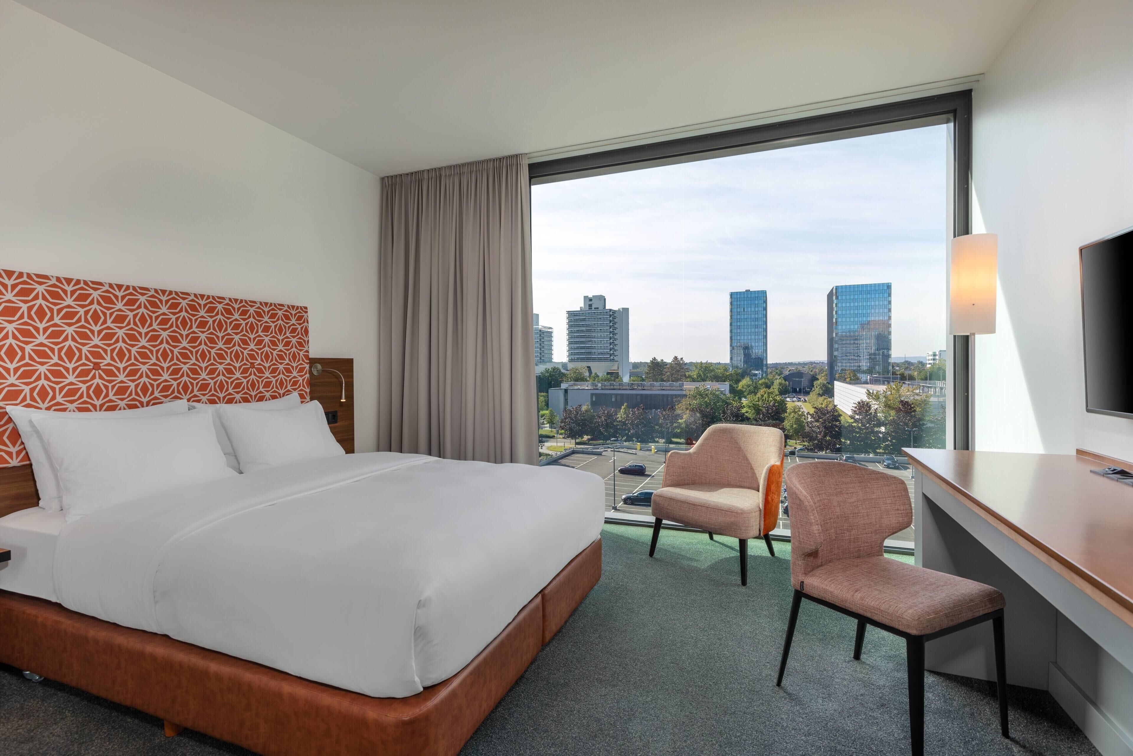Habitación de hotel con cama, escritorio, silla, televisión y un ventanal con vistas a la ciudad.