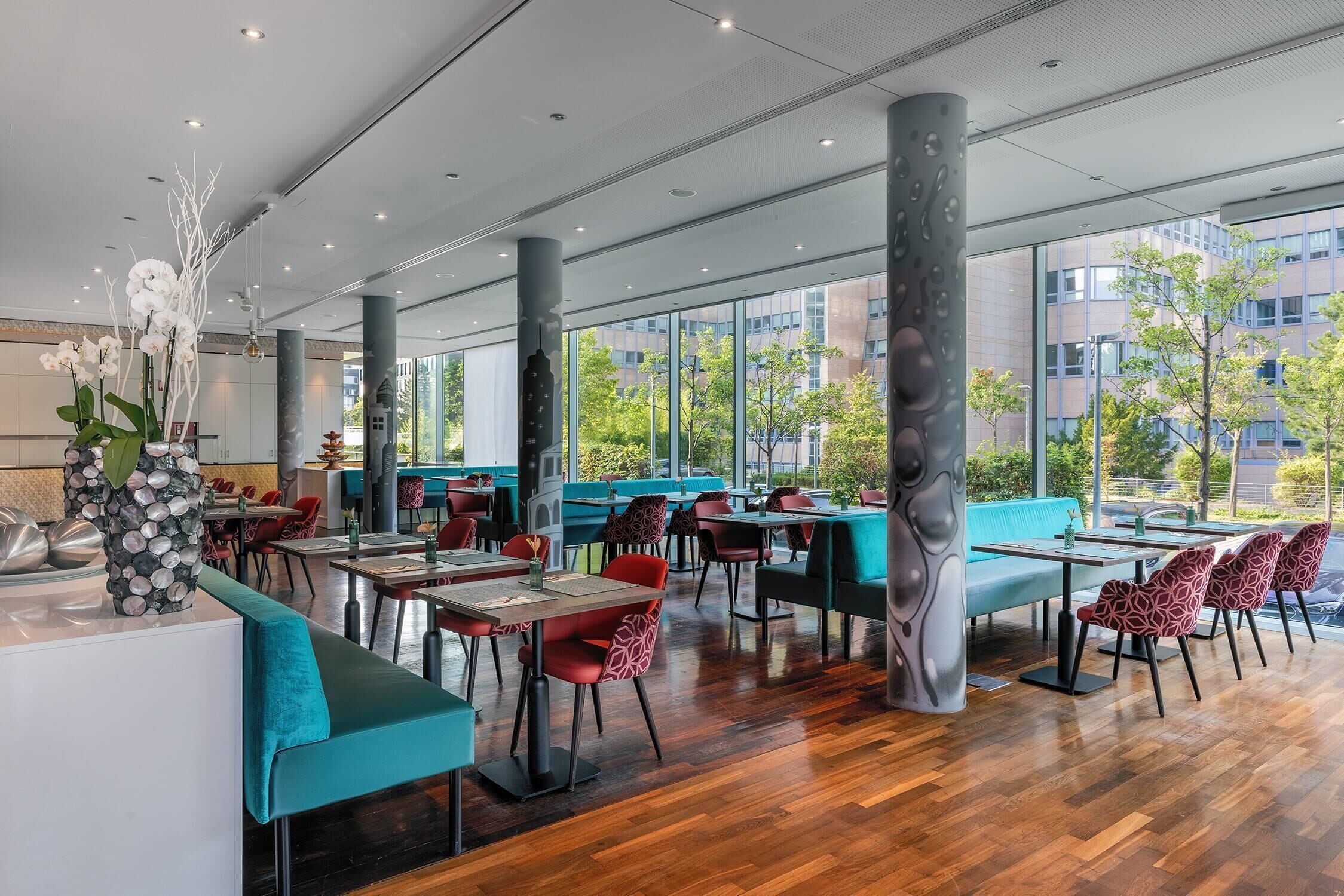 A modern restaurant with wooden flooring, teal and red seating, and large windows offering a view of the city.