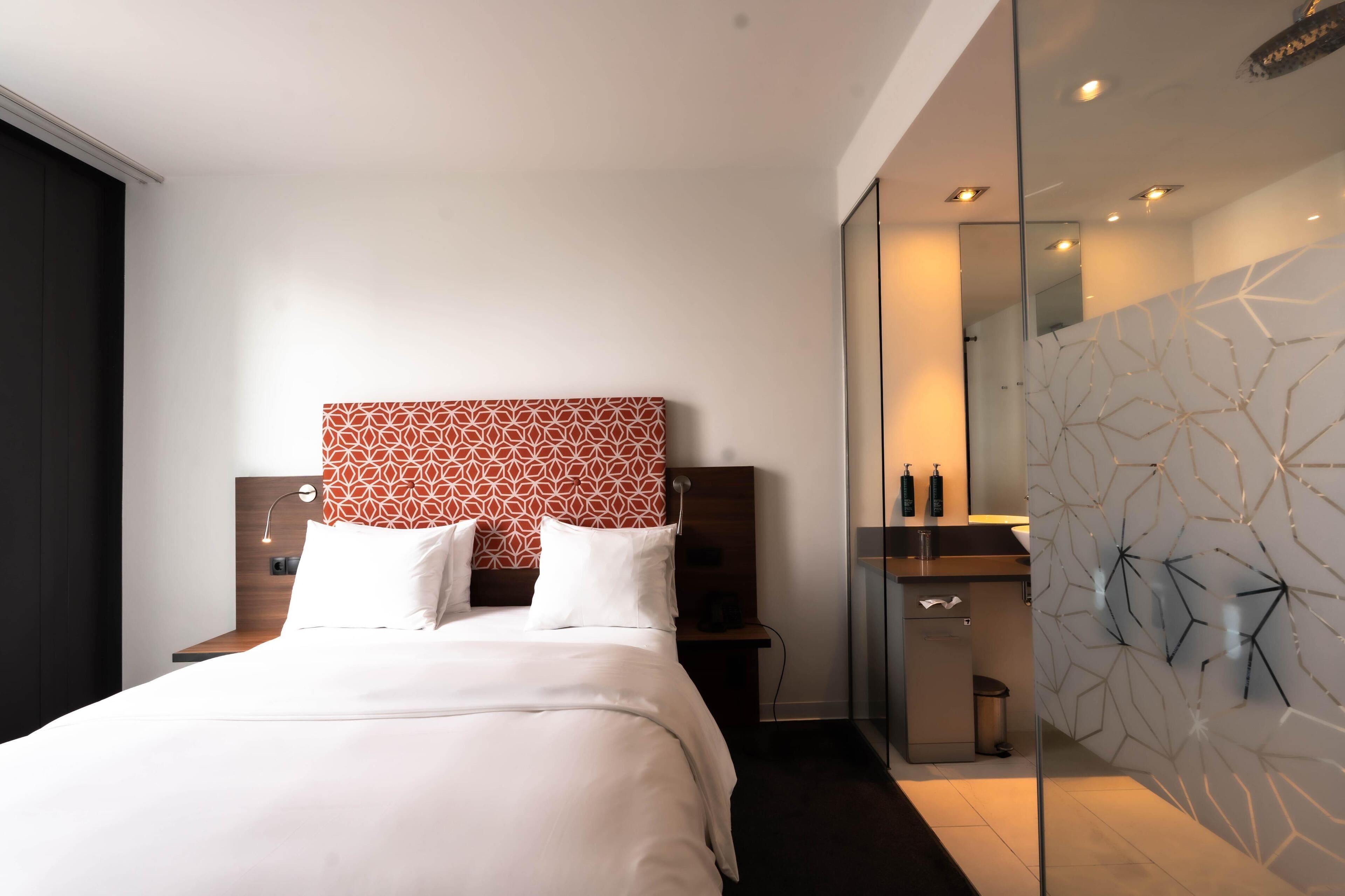 Habitación de hotel moderna con una cama grande, un separador de vidrio y un baño con diseño geométrico en la pared.
