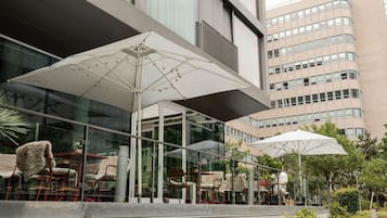 Un área de estar al aire libre con sombrillas y sillas, una pared de vidrio y un edificio al fondo.