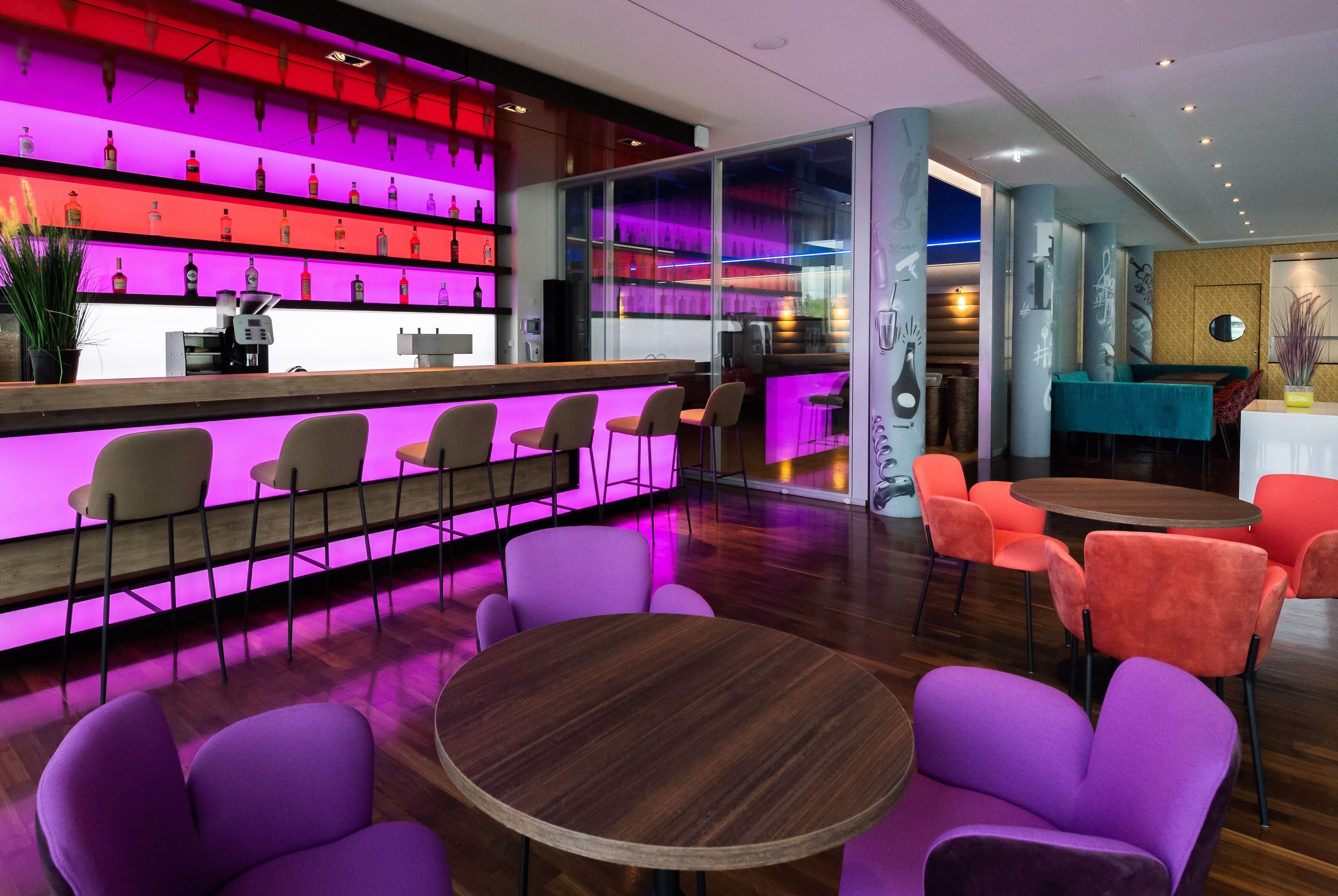 A bar area with a neon-lit shelf displaying bottles, a counter with a phone and a cash register, and seating with round tables and chairs.