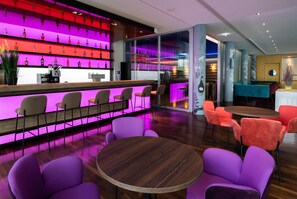Zona de bar con estante iluminado con neón que exhibe botellas, mostrador con teléfono y caja registradora, y asientos con mesas y sillas redondas.