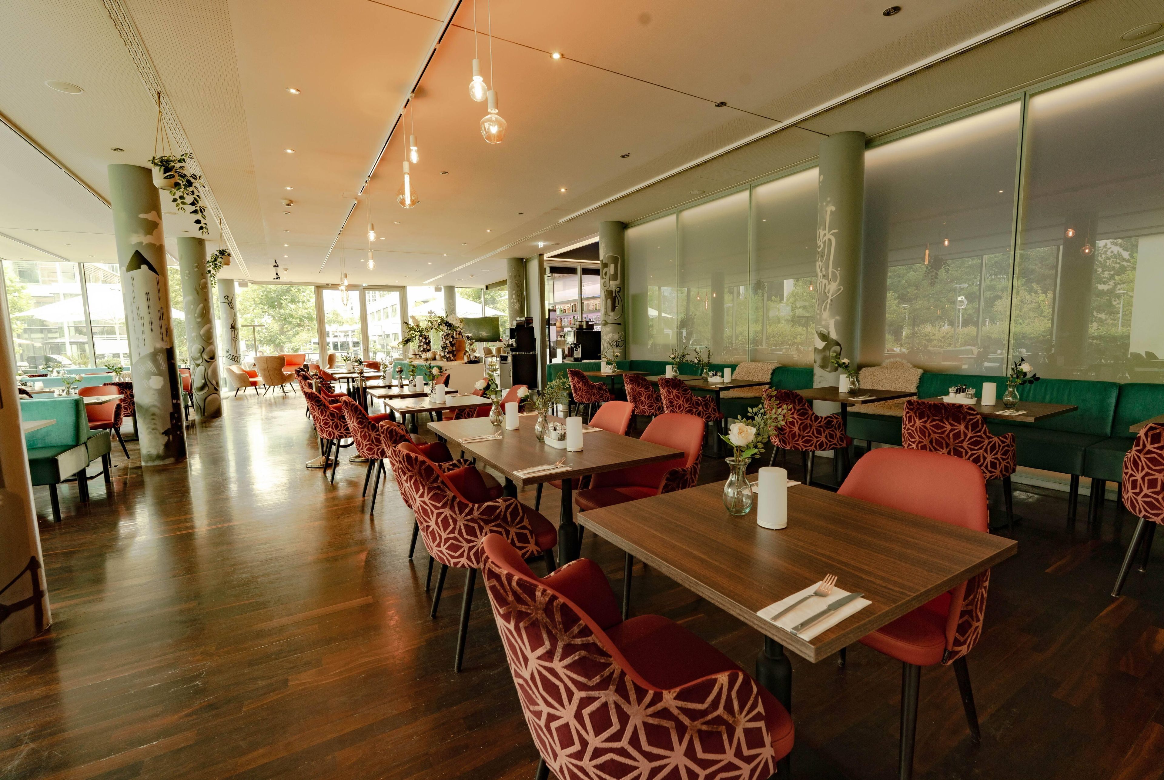 Un restaurante espacioso con mesas de madera y sillas estampadas, ventanas amplias que ofrecen vistas al exterior y lámparas colgantes.