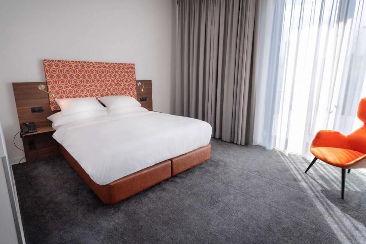 Habitación de hotel con cama, mesita de noche, una silla y ventana con cortinas.