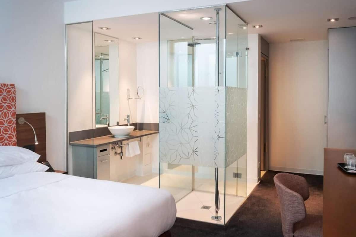 Una habitación de hotel moderna con una cama, un box de ducha con paredes de vidrio, un baño con lavamanos y un escritorio con silla.