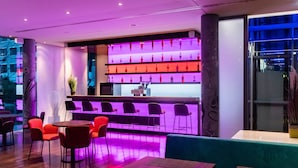 Zona de bar moderna con estantería iluminada en morado, sillas rojas y negras, y una barra.