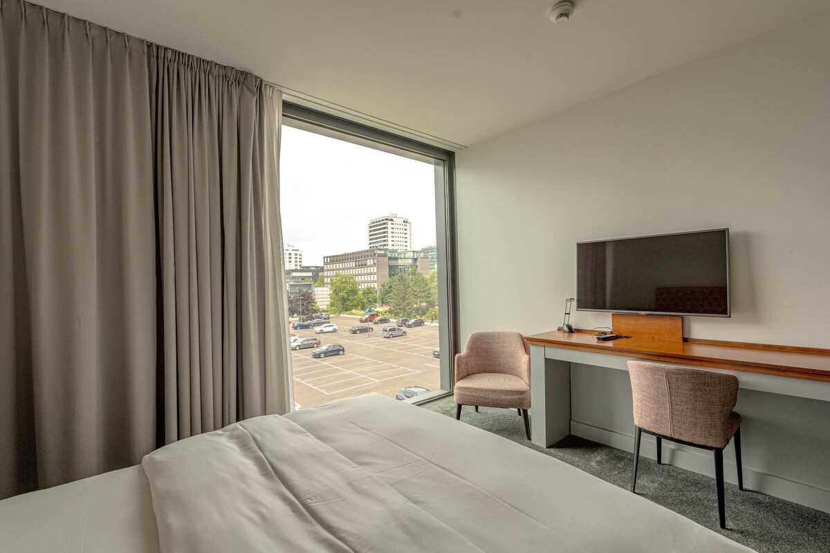 Habitación de hotel con cama, escritorio, dos sillas, televisión y vista a un estacionamiento y edificios.