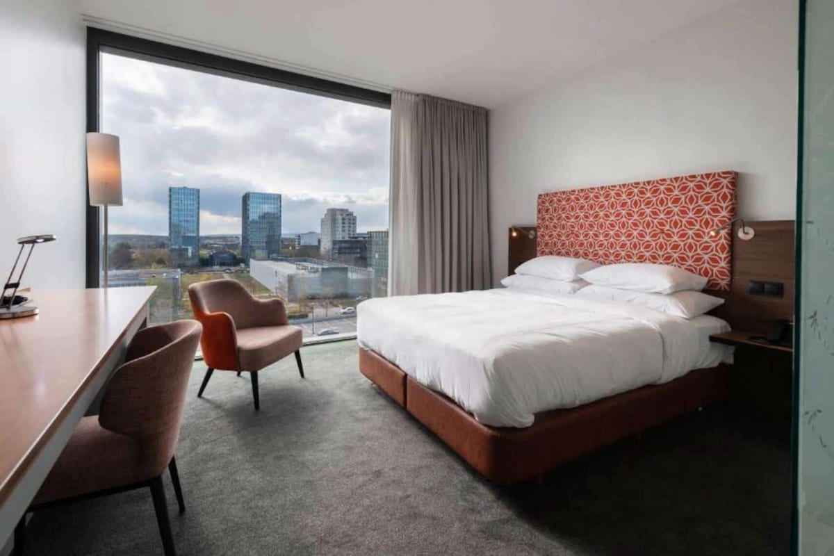 Habitación de hotel con cama, escritorio, dos sillas, un ventanal con vista a la ciudad y un cabecero estampado.