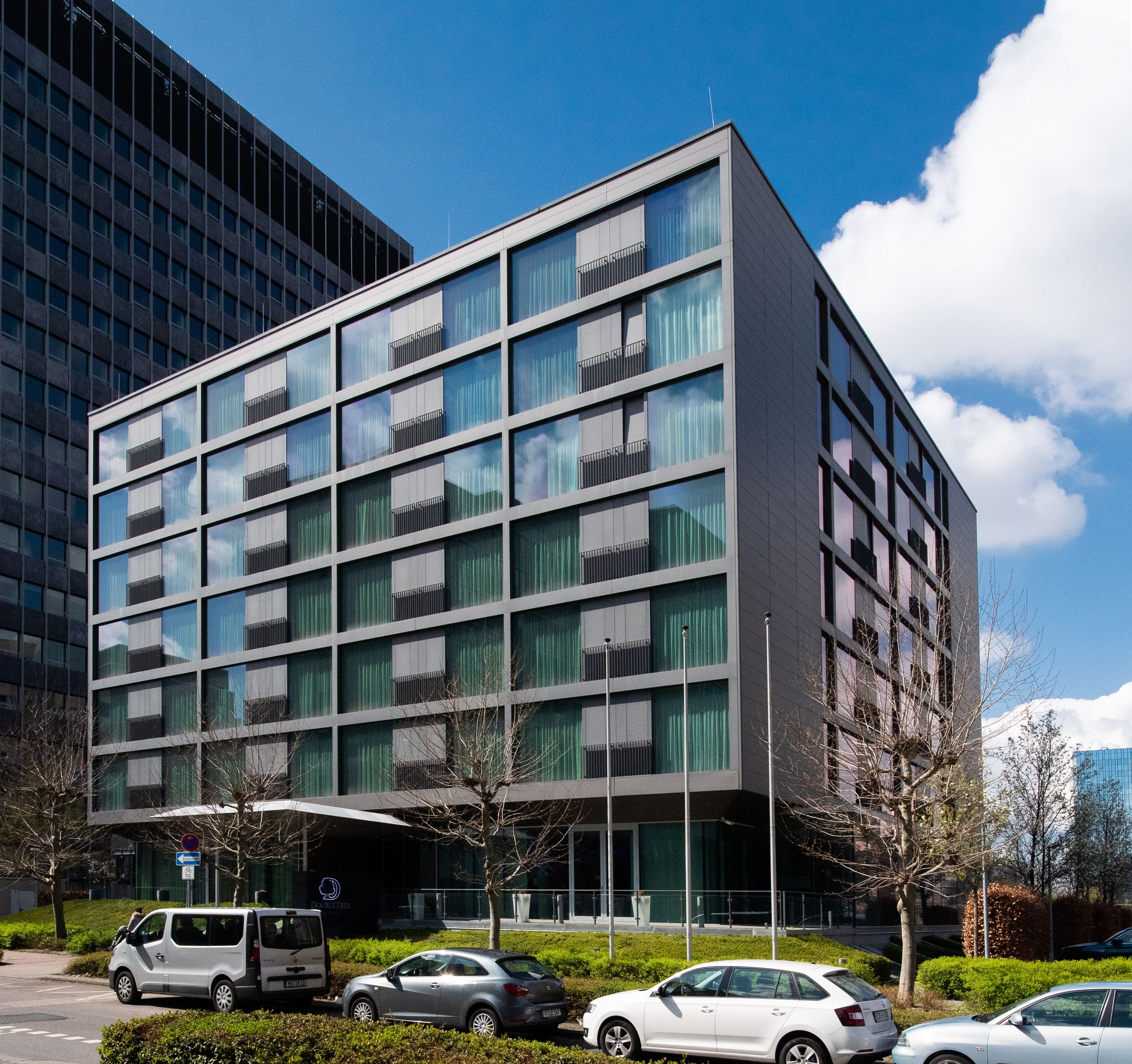 Un edificio moderno con una estructura de ventanas y balcones, rodeado de coches aparcados y vegetación.