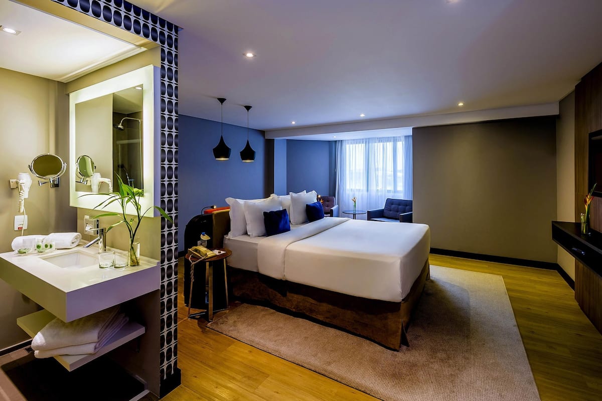 Ein modernes Hotelzimmer mit einem großen Bett, einem Badezimmer mit Spiegel und Waschbecken sowie einer Sitzecke mit Tisch und Stühlen.